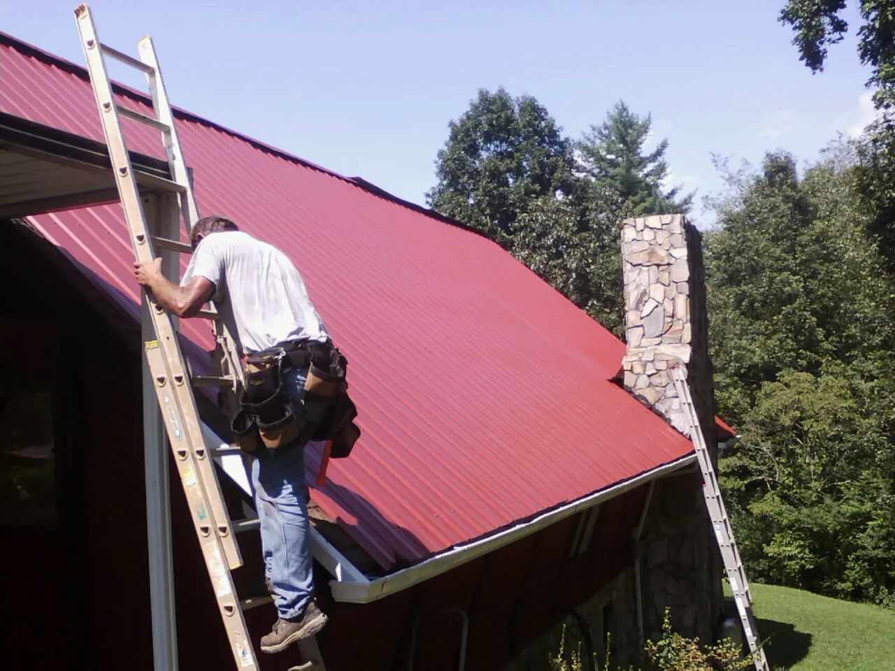 Roofer on a ladder beside a red metal roof and stone chimney — Soffit & Fascia Repair by Roofing Brothers in Church View
